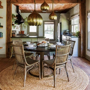 May include: A round jute rug with a woven pattern sits under a round wooden dining table with six woven chairs. Three gold pendant lights hang over the table. The table is set with plates, bowls, and a basket of bread.