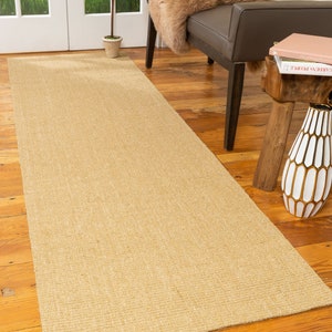 May include: A natural fiber runner rug in a light beige color. The rug is woven with a subtle texture and has a rectangular shape. It is placed on a wooden floor in a home setting.