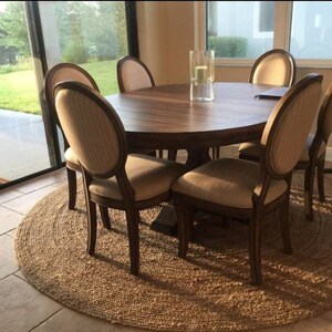 May include: A round wooden dining table with six chairs. The table has a natural wood finish and the chairs are upholstered in a light beige fabric. The table is set on a natural fiber rug.