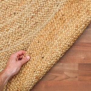 May include: A close-up of a natural fiber rug made of woven jute. The rug is light brown in color and has a textured surface. The rug is being rolled up, revealing the woven pattern.