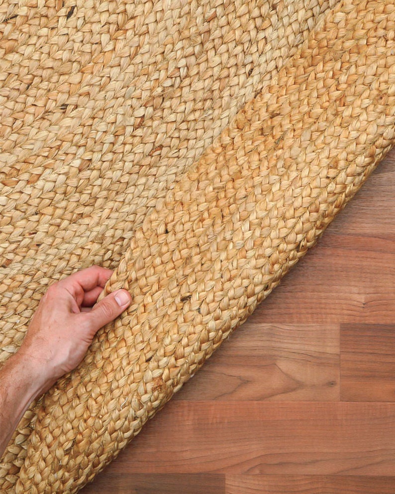 May include: A close-up of a natural fiber rug made of woven jute. The rug is light brown in color and has a textured surface. The rug is being rolled up, revealing the woven pattern.