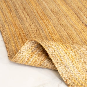 May include: A close-up of a natural fiber rug made from jute. The rug is a light brown color and has a woven texture. The rug is folded in half, showing the underside of the rug.