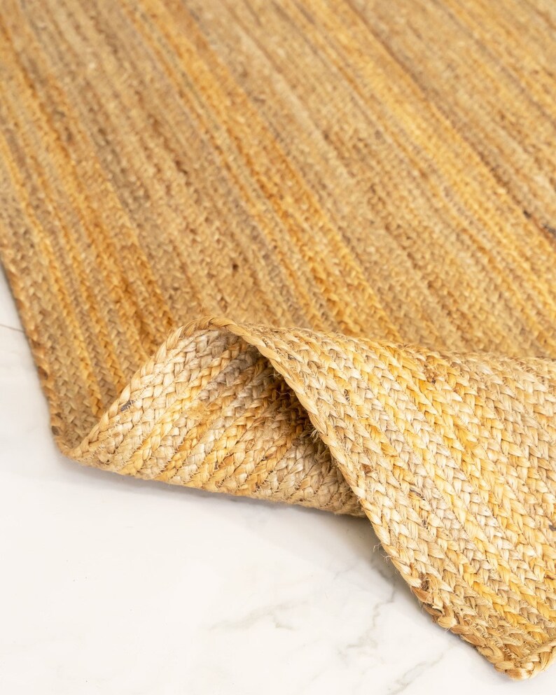 May include: A close-up of a natural fiber rug made from jute. The rug is a light brown color and has a woven texture. The rug is folded in half, showing the underside of the rug.