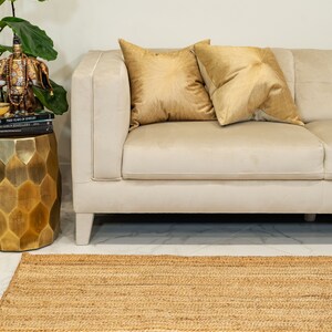May include: A light beige sofa with two gold throw pillows. The sofa is in front of a white wall and a gold decorative stand with a book on top. A natural fiber rug is in front of the sofa.