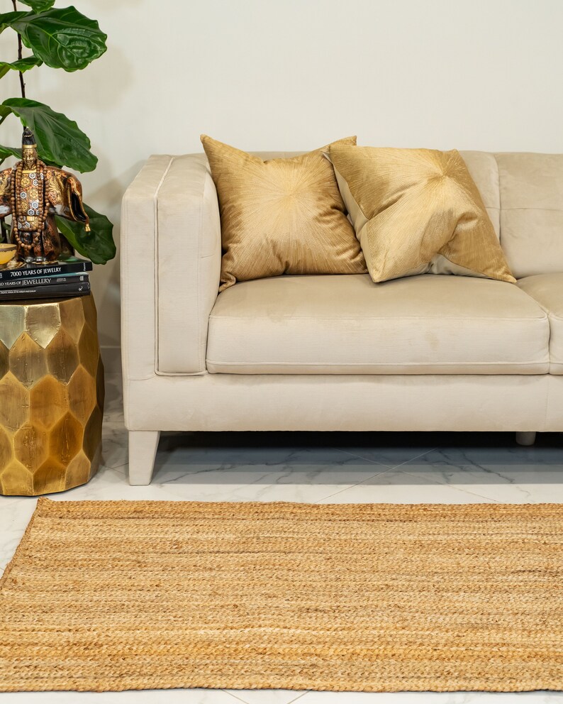 May include: A light beige sofa with two gold throw pillows. The sofa is in front of a white wall and a gold decorative stand with a book on top. A natural fiber rug is in front of the sofa.