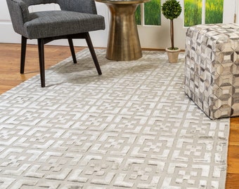 May include: A light gray area rug with a geometric pattern. The rug has a subtle sheen and is made of a soft, plush material. The rug is shown in a living room setting with a gray chair, a gold side table, and a patterned ottoman.