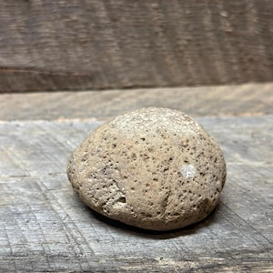 May include: A small, rounded, tan-colored stone with a porous, textured surface. The rock features numerous tiny indentations and pits across its exterior, giving it a weathered appearance. It has a slightly irregular, dome-like shape.
