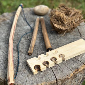 May include: A wooden fire-starting kit with a bow drill, a wooden board with carved notches, and a bundle of tinder. The bow drill has a blue and black cord. The kit is on a tree stump.