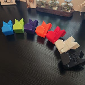 May include: A set of eight colorful plastic hooks with the text "Fence Buddy" printed on them. The hooks are arranged in a semi-circle on a black surface.