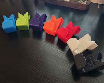 May include: A set of eight colourful plastic hooks with the text "Fence Buddy" printed on them. The hooks are arranged in a semi-circle on a black surface.