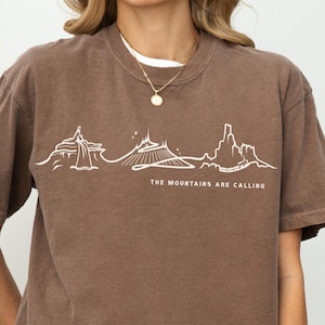 Może przedstawiać: Koszulka w kolorze espresso z białą grafiką gór i napisem "THE MOUNTAINS ARE CALLING". Koszulka ma krótkie rękawy i okrągły dekolt. Modelka ma na sobie białą koszulkę i czarne szorty.