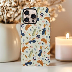 May include: A phone case with a light green background featuring a repeating pattern of croissants, bottles, blueberries, carrots, and cartoon mice. The case has a gold camera ring and a black border.