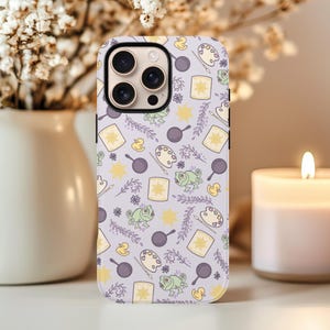 Puede incluir: Una funda de teléfono con un fondo lila claro y un patrón repetido de elementos de dibujos animados. El diseño incluye sartenes, paletas de pintura, ranas, patos y soles en amarillo, morado y verde.