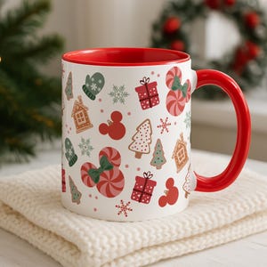 Puede incluir: Una taza de cer&aacute;mica blanca con interior y asa rojos, decorada con ilustraciones navide&ntilde;as. El dise&ntilde;o incluye casas de jengibre, bastones de caramelo, copos de nieve y cajas de regalo. La taza est&aacute; sobre una superficie de punto color crema.