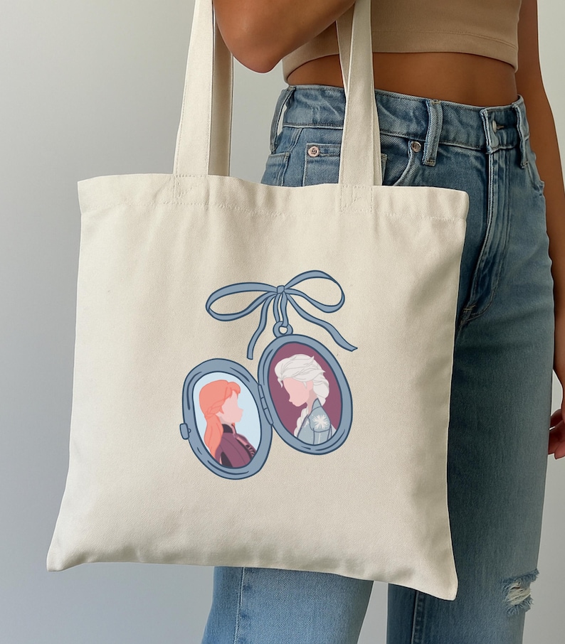May include: Cream-colored tote bag featuring a blue ribbon and two oval lockets. Each locket contains a profile illustration of a woman. One has red hair and the other has white hair. The bag is held by a person wearing blue jeans.
