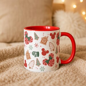 Puede incluir: Taza de cer&aacute;mica blanca con interior y asa rojos, adornada con un motivo festivo. El dise&ntilde;o incluye casas de jengibre, &aacute;rboles de Navidad, bastones de caramelo, copos de nieve y siluetas de Mickey Mouse en rojo, verde y blanco.