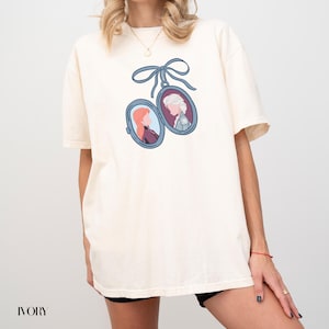Op de afbeelding: Ivoor kleurig t-shirt met een blauw lint en twee ovale medaillons met cartoonportretten. E&eacute;n medaillon toont een vrouw met rood haar, de andere een vrouw met wit haar. Het woord "IVORY" staat onderaan gedrukt.