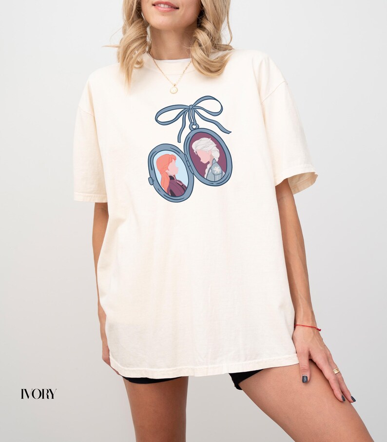 Op de afbeelding: Ivoor kleurig t-shirt met een blauw lint en twee ovale medaillons met cartoonportretten. E&eacute;n medaillon toont een vrouw met rood haar, de andere een vrouw met wit haar. Het woord "IVORY" staat onderaan gedrukt.
