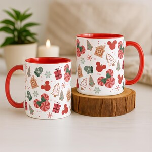Puede incluir: Dos tazas de cer&aacute;mica blanca con interiores y asas rojas. Las tazas est&aacute;n decoradas con un patr&oacute;n festivo de bastones de caramelo, casas de jengibre, &aacute;rboles de Navidad y siluetas de Mickey Mouse. Una taza est&aacute; sobre un posavasos de madera.