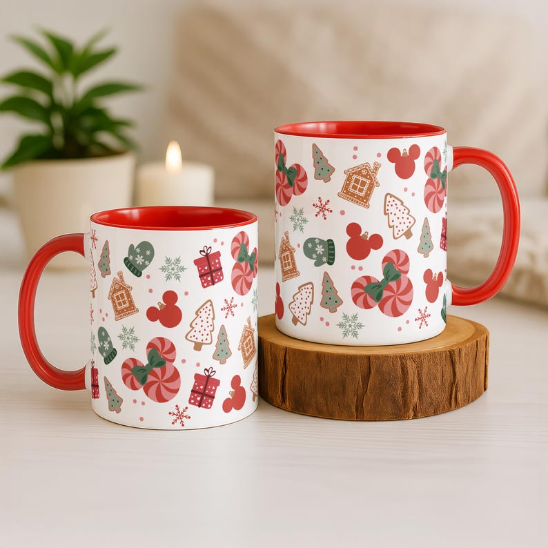Puede incluir: Dos tazas de cer&aacute;mica blanca con interiores y asas rojas. Las tazas est&aacute;n decoradas con un patr&oacute;n festivo de bastones de caramelo, casas de jengibre, &aacute;rboles de Navidad y siluetas de Mickey Mouse. Una taza est&aacute; sobre un posavasos de madera.