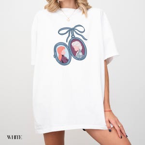 Op de afbeelding: Wit T-shirt met een blauw lint en twee ovale medaillons, elk met een cartoonportret van een vrouw. De portretten staan in blauwe kaders. Het woord "WHITE" staat onderaan gedrukt.