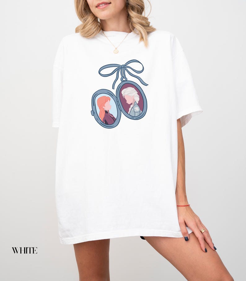 Op de afbeelding: Wit T-shirt met een blauw lint en twee ovale medaillons, elk met een cartoonportret van een vrouw. De portretten staan in blauwe kaders. Het woord "WHITE" staat onderaan gedrukt.