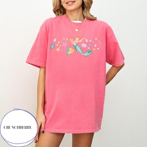 May include: Pink t-shirt featuring a colorful illustration of a mermaid, fish, and various sea elements. The design includes a mermaid with red hair and a green tail, along with other aquatic details. The shirt has short sleeves and a round neckline.