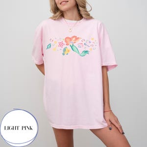May include: Light pink t-shirt featuring a colorful mermaid graphic. The design includes a mermaid, starfish, and other aquatic elements. The shirt has the text "LIGHT PINK" in a circle at the bottom.