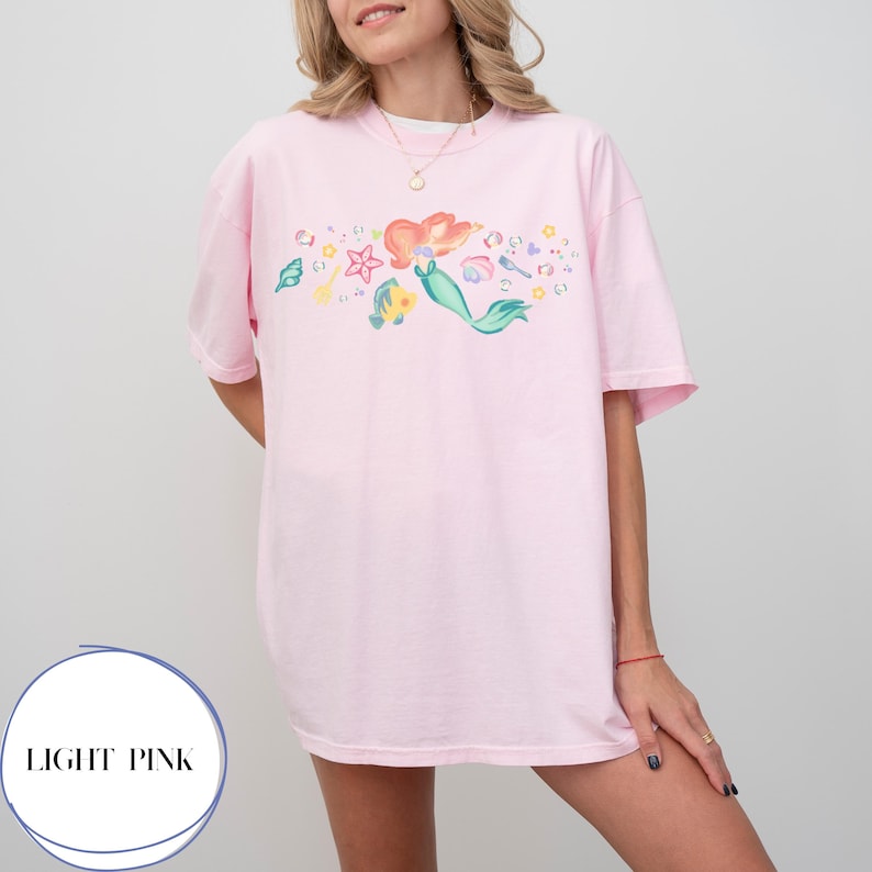 May include: Light pink t-shirt featuring a colorful mermaid graphic. The design includes a mermaid, starfish, and other aquatic elements. The shirt has the text "LIGHT PINK" in a circle at the bottom.