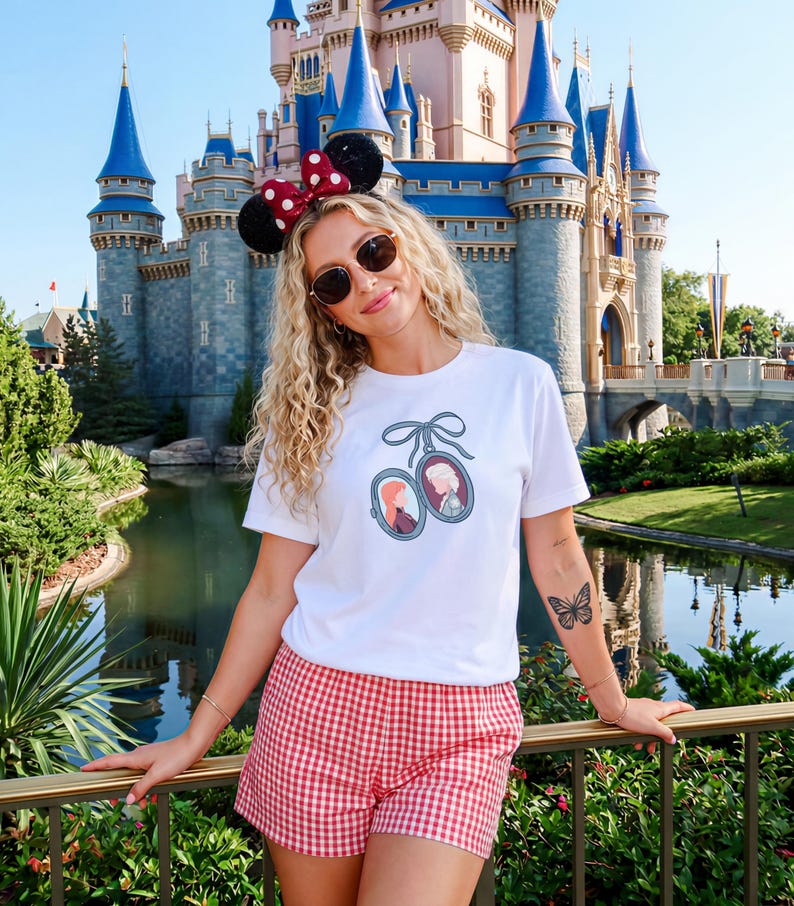 Op de afbeelding: Een vrouw draagt een wit t-shirt met een ingelijst portretontwerp, rood-witte geruite shorts en een Minnie Mouse oren-hoofdband. Ze draagt een zonnebril en heeft een vlinder-tattoo op haar arm. Een kasteel is op de achtergrond.