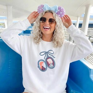 Op de afbeelding: Wit sweatshirt met een cartoon illustratie van twee karakters in medaillons. De persoon draagt een lichtblauwe strik hoofdband met paarse oren en witte stippen en een zonnebril.