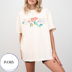 May include: Ivory-colored t-shirt featuring a colorful illustration of a mermaid with red hair and a green tail, surrounded by aquatic elements. The shirt has short sleeves and a relaxed fit. The word "IVORY" is printed in a circle at the bottom.