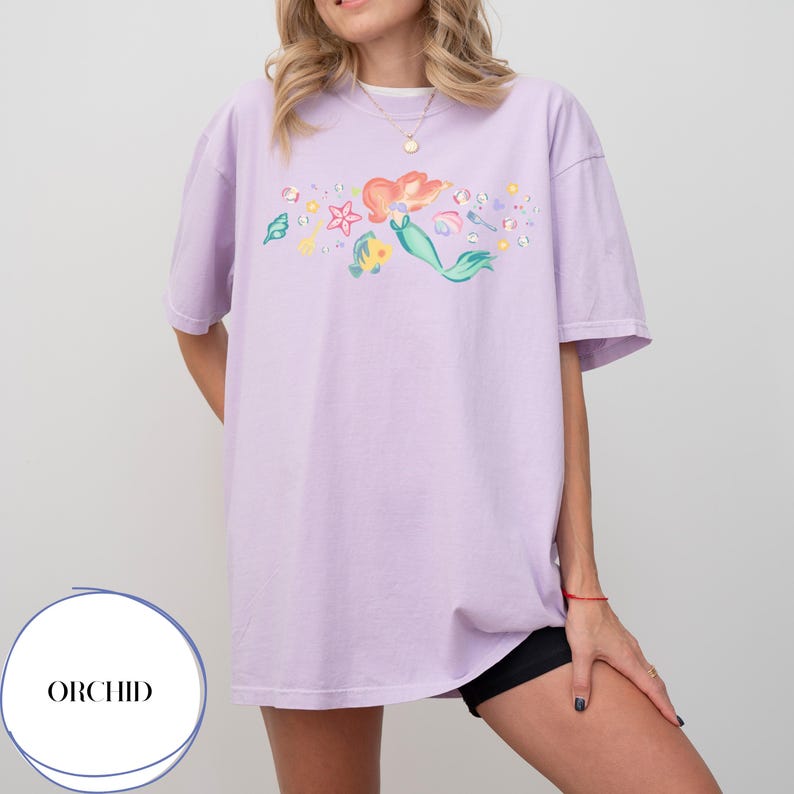 May include: A light purple t-shirt featuring a colorful illustration of a mermaid with red hair and a green tail, surrounded by sea creatures and bubbles. The shirt has short sleeves and a relaxed fit. The word "ORCHID" is in a white circle in the lower left corner.