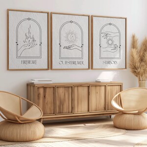 May include: Three minimalist black and white prints with arched frames and hand drawn illustrations. The prints are titled "Fireheart", "Cursebreaker", and "Starborn". The prints are hanging on a white wall above a wooden cabinet.