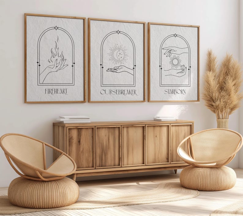 May include: Three minimalist black and white prints with arched frames and hand drawn illustrations. The prints are titled "Fireheart", "Cursebreaker", and "Starborn". The prints are hanging on a white wall above a wooden cabinet.