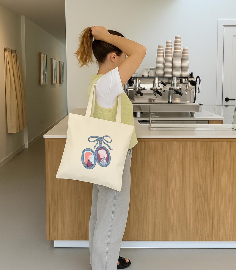May include: Cream-colored tote bag featuring a blue oval locket design with two portraits. The locket is tied with a blue bow. The bag has a simple, rectangular shape with two handles.