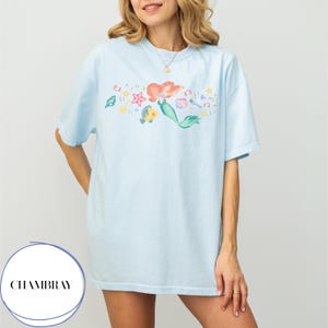 May include: Light blue t-shirt featuring a colorful illustration of a mermaid with red hair and a green tail, along with various sea creatures. The shirt has a relaxed fit and is made of soft material. The brand name "CHAMBRAY" is printed on the bottom left.