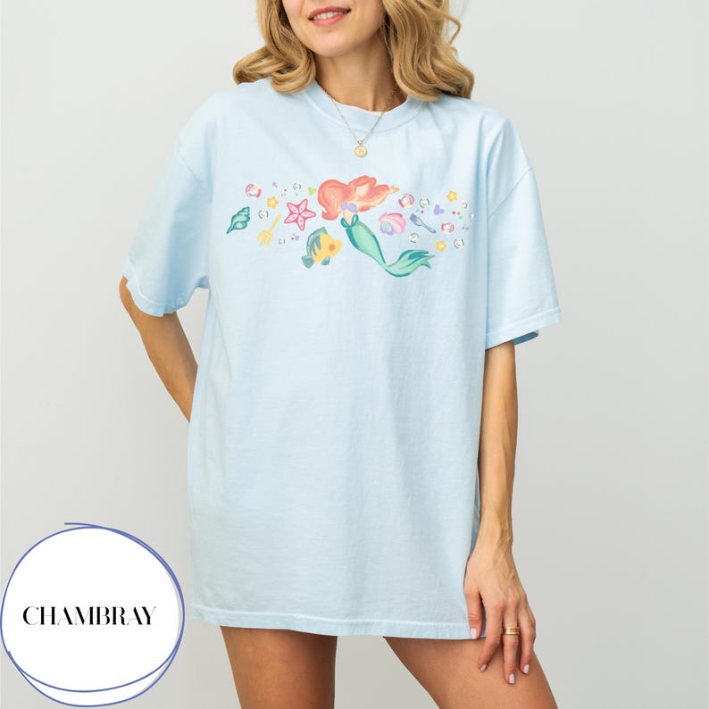 May include: Light blue t-shirt featuring a colorful illustration of a mermaid with red hair and a green tail, along with various sea creatures. The shirt has a relaxed fit and is made of soft material. The brand name "CHAMBRAY" is printed on the bottom left.