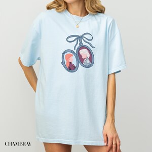 Op de afbeelding: Lichtblauw T-shirt met een hangerontwerp met twee portretten van animatiefiguren, opgehangen aan een blauw lint. Het woord "CHAMBRAY" staat linksonder op het shirt.