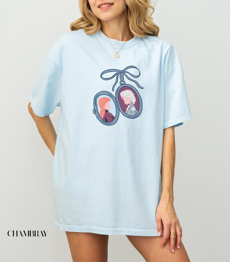 Op de afbeelding: Lichtblauw T-shirt met een hangerontwerp met twee portretten van animatiefiguren, opgehangen aan een blauw lint. Het woord "CHAMBRAY" staat linksonder op het shirt.