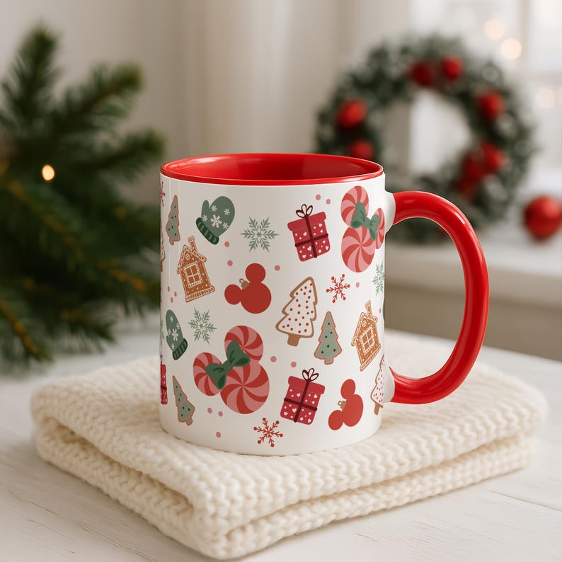 Puede incluir: Una taza de cer&aacute;mica blanca con interior y asa rojos, decorada con ilustraciones navide&ntilde;as. El dise&ntilde;o incluye casas de jengibre, bastones de caramelo, copos de nieve y cajas de regalo. La taza est&aacute; sobre una superficie de punto color crema.