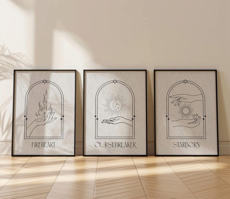 May include: Three black framed prints with white backgrounds and black line drawings. The first print features a hand holding a flame and the text "Fireheart". The second print features a hand holding a crescent moon and stars and the text "Cursebreaker". The third print features a hand holding a sun and the text "Starborn".