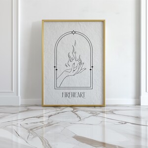 May include: A black and white line art print of a hand holding a flame with the word "FIREHEART" below it. The print is framed in a gold frame and is hanging on a white wall.