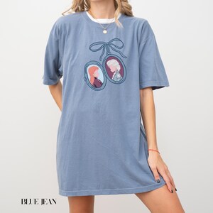 Op de afbeelding: Blauw spijkerbroek T-shirt met een dubbel ovaal hangerontwerp met cartoonportretten, verbonden door een blauw lint. Het shirt heeft een effen blauwe kleur en korte mouwen. De tekst "BLUE JEAN" staat onderaan.