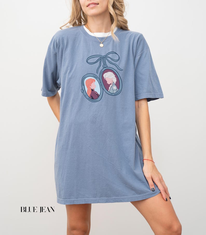 Op de afbeelding: Blauw spijkerbroek T-shirt met een dubbel ovaal hangerontwerp met cartoonportretten, verbonden door een blauw lint. Het shirt heeft een effen blauwe kleur en korte mouwen. De tekst "BLUE JEAN" staat onderaan.