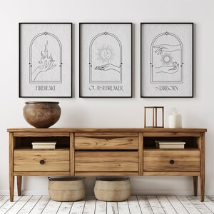 May include: Three black framed prints with white backgrounds and black line drawings of hands holding a flame, a crescent moon, and a sun. The prints are titled "Fireheart", "Cursebreaker", and "Starborn".