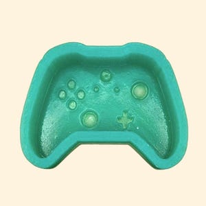 May include: A teal-colored, molded plastic controller-shaped tray. The tray features the button layout of a gaming controller, including a directional pad, buttons, and analog sticks. The tray is designed to hold small items.