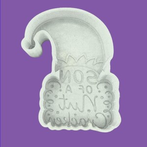 May include: A white plastic cookie cutter shaped like a curved elf hat with a rectangular base. The interior of the base features embossed text that reads SON OF A NUTCRACKER in a stylized script font, surrounded by decorative scrollwork.