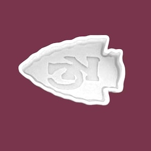 May include: A white arrowhead-shaped design with the letters "KC" inside. The design is raised and has a textured appearance. The background is a solid maroon color.