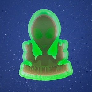 May include: A neon green alien figure with a hooded design and large eyes, set against a dark blue starry background. The figure is holding up two fingers and the text "HOME IN DEEP" is visible.